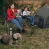 Två personer och hund vid campingplats med campingstolar och tält.
