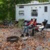 Två personer i campingstolar framför husvagn i skog.